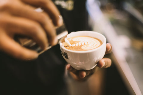 Barista gießt geschäumte Milch in eine Cappuccino Tasse und gestaltet kunstvolle Latte Art mit Herz- und Blattmuster.
