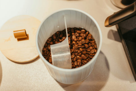 Kaffeebohnen in einem offenen Behälter, von oben fotografiert. Im Behälter liegt ein kleines Schäufelchen. Neben dem Behälter der Deckel