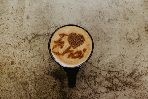Tasse mit Chai Latte von oben fotografiert, steht auf einem grauen Untergrund. Im Schaum des Lattes steht "I love Chai" mit Kakao Pulver