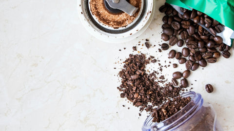 frisch gemahlener, grober Kaffee neben Kaffeebohnen und einer geöffneten Kaffeemühle auf einer hellen Arbeitsfläche